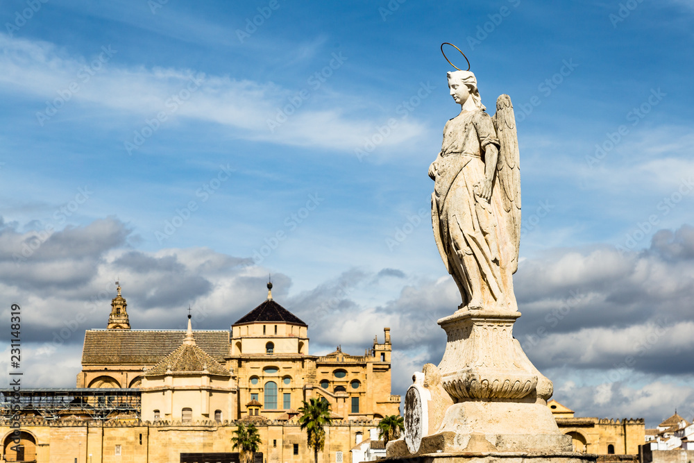 View of Mezquita, Catedral de Cordoba behind an angel statue on the roman bridge. A former Moorish Mosque that is now the Cathedral of Cordoba, Mezquita is a UNESCO World Heritage Site.