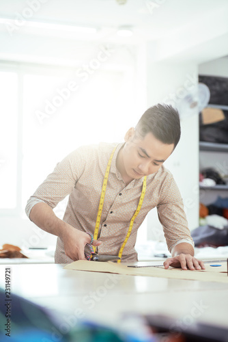Concentrated young male tailor working at his workplace, he cutting material with scissors