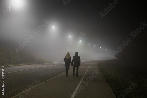 Young couple holding each other hands. Slowly walking under white street lights in night. Dark time. Peaceful atmosphere in mist. Foggy air. © fotoduets