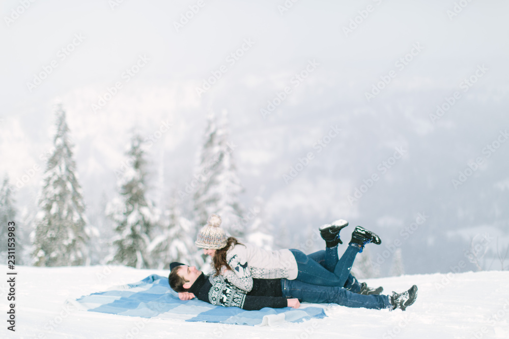 Charming young couple in warm winter knitted sweaters lying on the wool blue chekered blanket in snowy forest. Love in winter, Happy New Year and Christmas concept Charming young couple in warm winter knitted sweaters lying on the wool blue chekered blanket in snowy forest. Love in winter, Happy New Year and Christmas concept