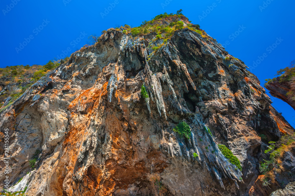 Obraz premium Look up at the sheer hanging orange brown limestone cliff like stone icicles hanging above people bathing in the sea. Phra Nang Princess Cave Beach, Krabi, Thailand.
