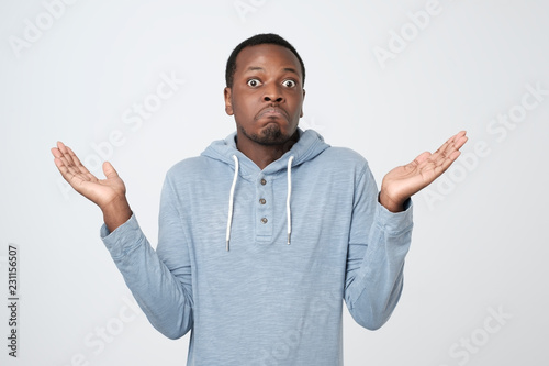 Young african man in blue shirt being at a loss, showing helpless gesture with arm and hand