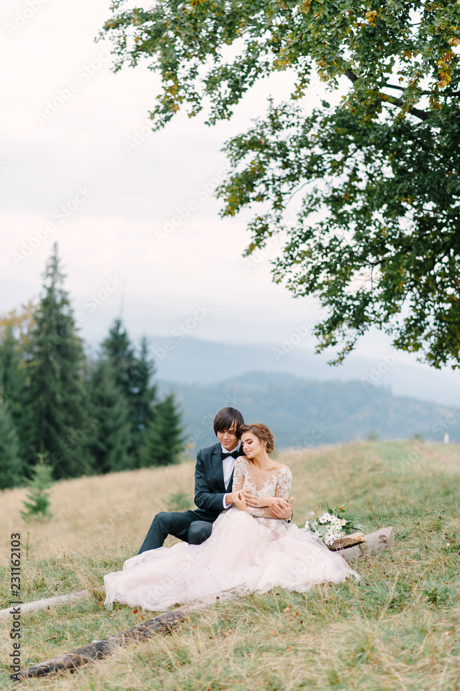 Attractive couple newlyweds, happy and joyful moment. Man and woman in festive clothes sit on the stones near the wedding decoration in boho style. Ceremony outdoors.