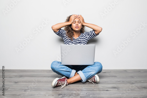 Фотография Sad skinned curly woman dressed in casual outfit, sits bare foot on wooden floor in empty room with laptop
