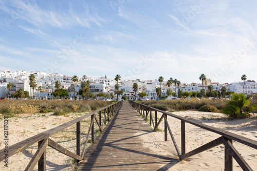 Wooden boardwalk leads between palm trees to the white city of Conil de la Frontera in Andalusia with blue sky