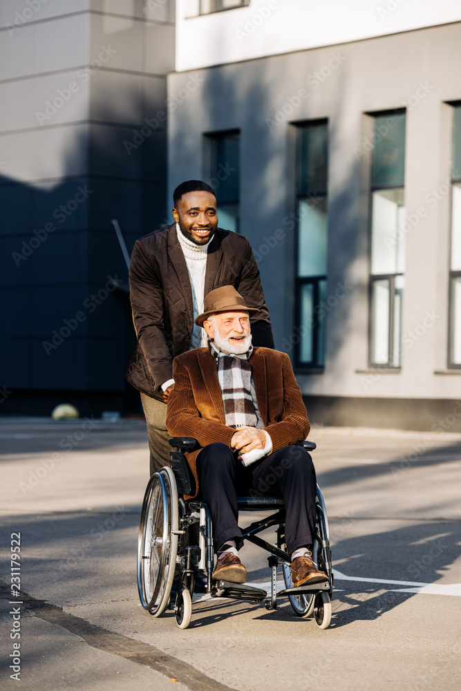 smiling senior disabled man in wheelchair and african american cuidador riding by street