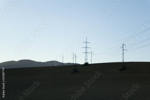 Electricity pylon - pole for transmission of electricity and electric energy. Minimalist nature of field and hills around facility. Dark evening with dark ground