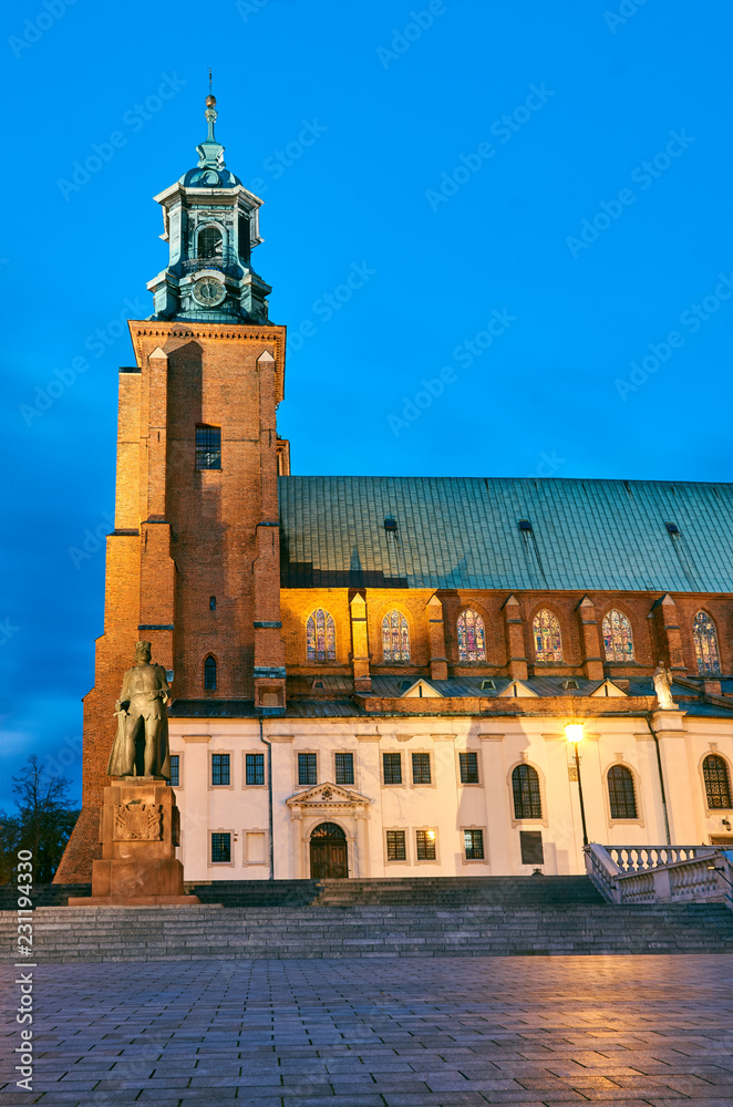 Statue and Gothic cathedral church by night in Gniezno .