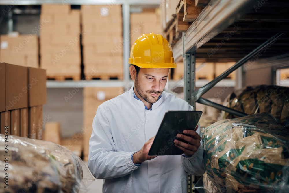 Young Caucasian worker using tablet while standing in warehouse. On head helmet. Young Caucasian worker using tablet while standing in warehouse. On head helmet.