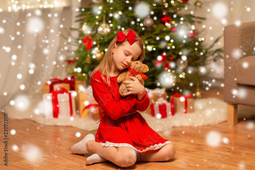 christmas, holidays and childhood concept - girl in red dress hugging teddy bear at home