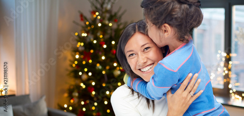 family, holidays and happiness concept - happy mother hugging her daughter over christmas thee lights background