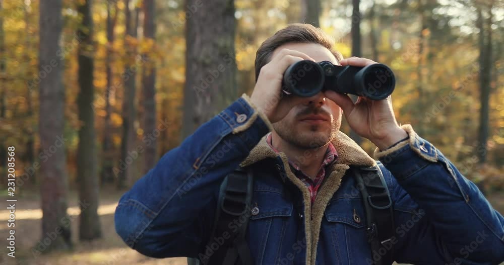 Close up of the Caucasian male tourist with backpack looking in the binoculars while standing in the forest at fall. Outdoor. Close up of the Caucasian male tourist with backpack looking in the binoculars while standing in the forest at fall. Outdoor.