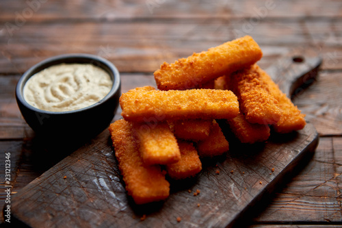 Fotografie Pile of golden fried fish fingers with white garlic sauce placed on chopping board on dark wooden background