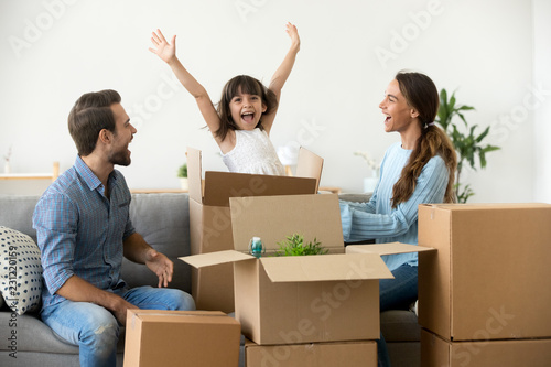 Multi-ethnic family sitting on couch in living room funny daughter jumping out of carton box parents laughing play with child have fun together move at new house. Mortgage loan and relocating concept