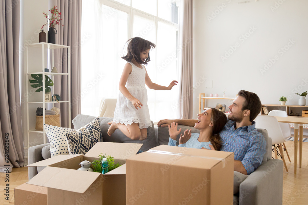 Multi-ethnic happy mother father sitting in living room looking at little child have fun jumping on sofa. People surrounded by cardboard boxes full with belongings. Moving at new home or loan concept Multi-ethnic happy mother father sitting in living room looking at little child have fun jumping on sofa. People surrounded by cardboard boxes full with belongings. Moving at new home or loan concept