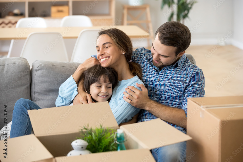 Multi-ethnic happy family sitting together on couch in living room. Loving mother embrace little adorable preschool daughter spending free time at new home. Moving relocating or mortgage loan concept