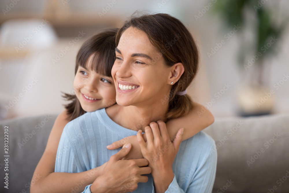 Head shot portrait smiling attractive female sitting embracing with little adorable daughter on couch in living room at home looking away. Happy motherhood new mother for adopted child concept Head shot portrait smiling attractive female sitting embracing with little adorable daughter on couch in living room at home looking away. Happy motherhood new mother for adopted child concept