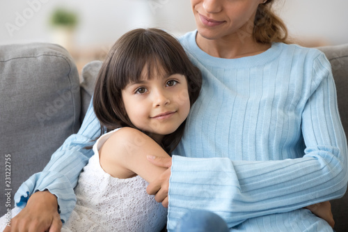 Close up portrait beautiful little girl sitting on sofa hugging mommy looking at camera. Loving single mother embrace small daughter showing love and support. Mum kid sincere warm relationship concept