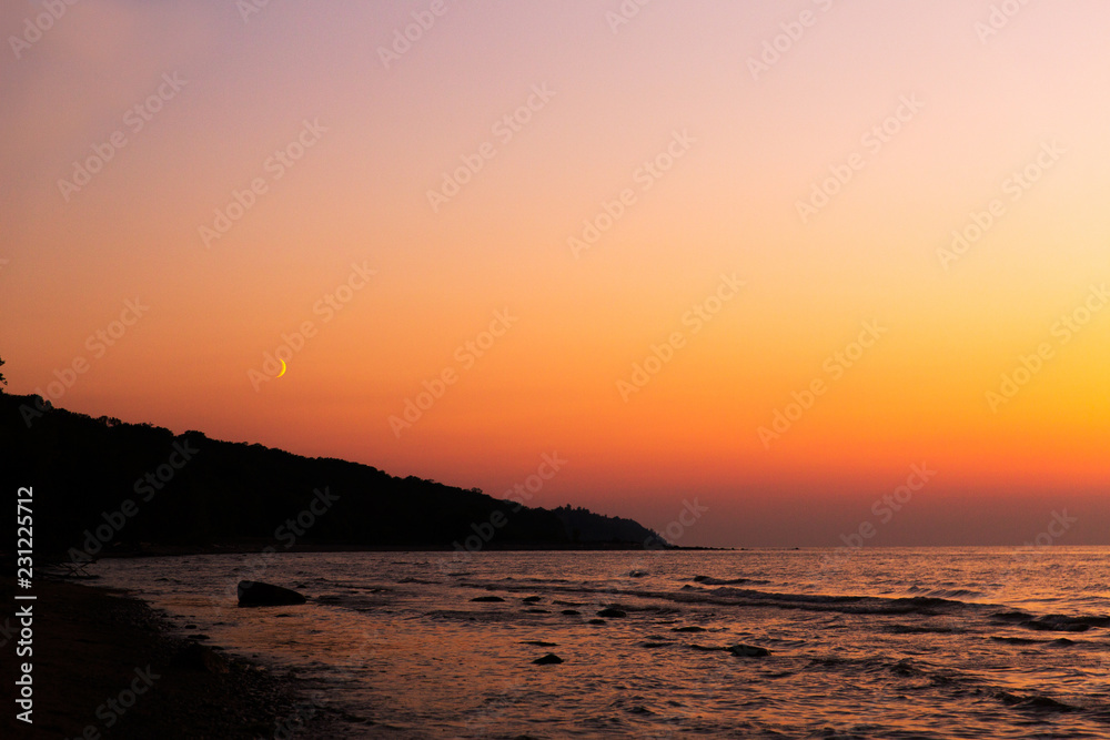 Moon in the sky. Sunset on the sea. Beautiful evening landscape. Silhouette of forest on the sea coast