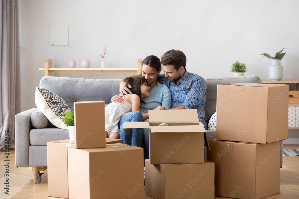 Diverse family parents and little daughter sitting together on sofa in living room at new home. People surrounded by carton boxes resting indoors. Buy new house and improve living conditions concept Diverse family parents and little daughter sitting together on sofa in living room at new home. People surrounded by carton boxes resting indoors. Buy new house and improve living conditions concept
