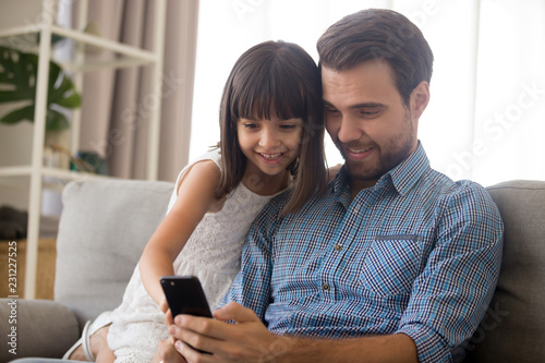 Cheerful positive multi-ethnic family sitting on couch in living room at home Caucasian father daughter smiling looking on smartphone screen have fun watching cartoons chatting playing games.
