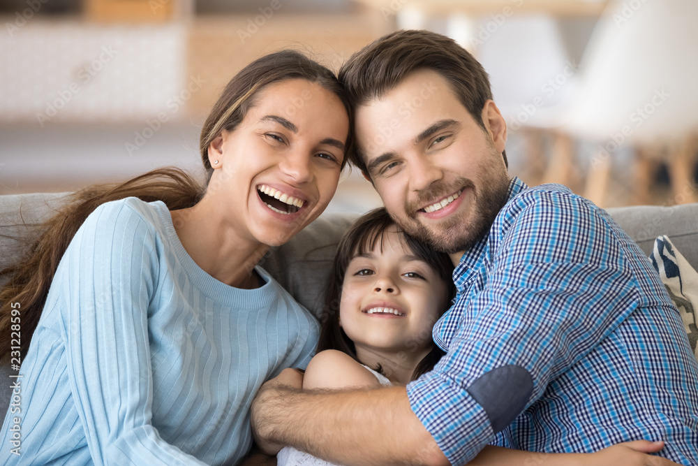 Head shot portrait happy diverse cheerful laughing multi-ethnic family with little adorable sweet daughter embracing sitting on couch in living room enjoying free time together at home on weekend. Head shot portrait happy diverse cheerful laughing multi-ethnic family with little adorable sweet daughter embracing sitting on couch in living room enjoying free time together at home on weekend.