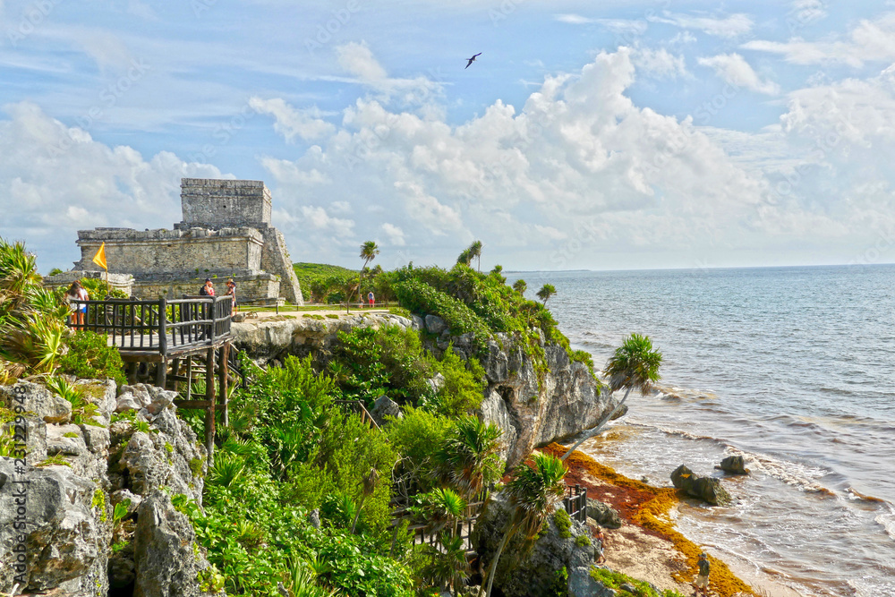 Tulum, Quintana Roo / Mexico - August 2018: The Castle of Tulum with a bird flying over it, some tourists in the viewpoint and the beach full of sargasso algae.