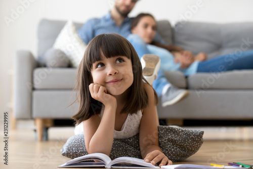 Close up little adorable thoughtful smiling daughter dreaming lying at cushion on warm floor with book in living room at modern home, resting married couple parents on background, focus on small kid