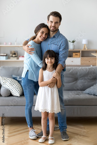 Full length diverse multi-ethnic family married couple wife husband little daughter embracing standing together in living room smiling looking at camera at new modern home feels happy and satisfied.