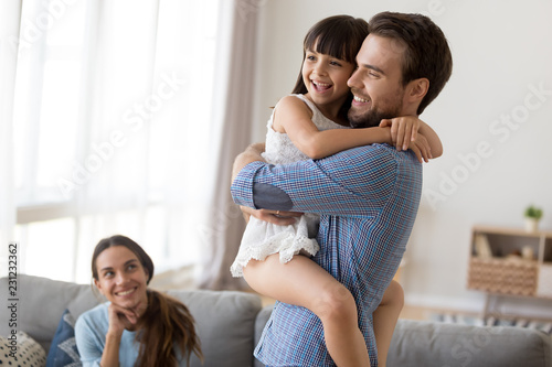Multi-ethnic couple and kid in living room at home. Laughing young father hold on hands embrace little smiling daughter, mother sitting on couch looking at beloved people. Happy diverse family concept photo
