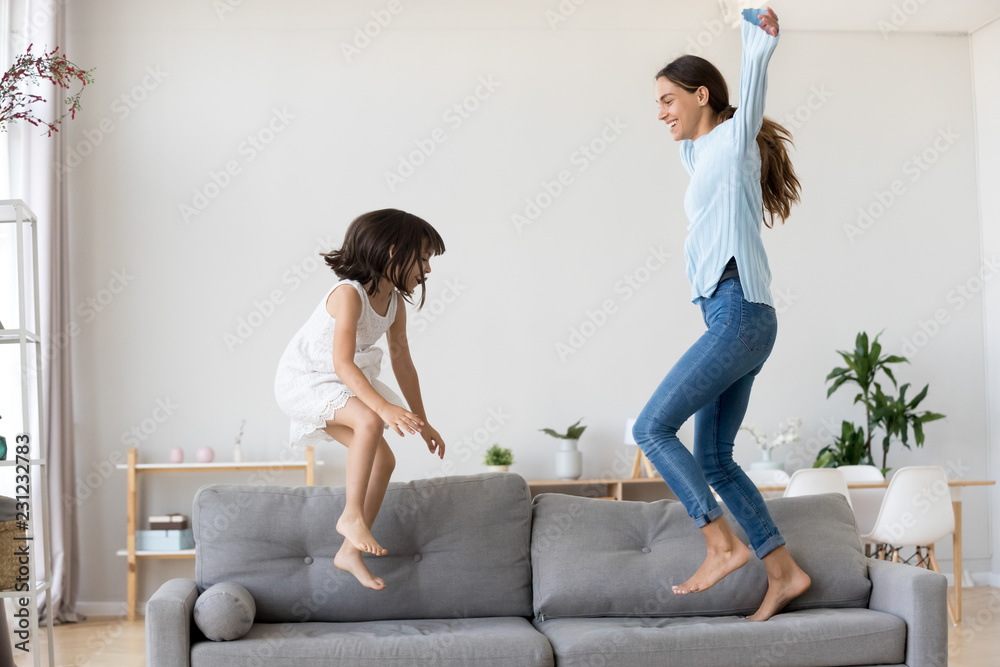 Full length positive mother and little daughter having fun jumping together on sofa in living room at home. Adorable girl playing enjoying active weekends with elder sister nanny or loving mom indoors - obrazy, fototapety, plakaty 