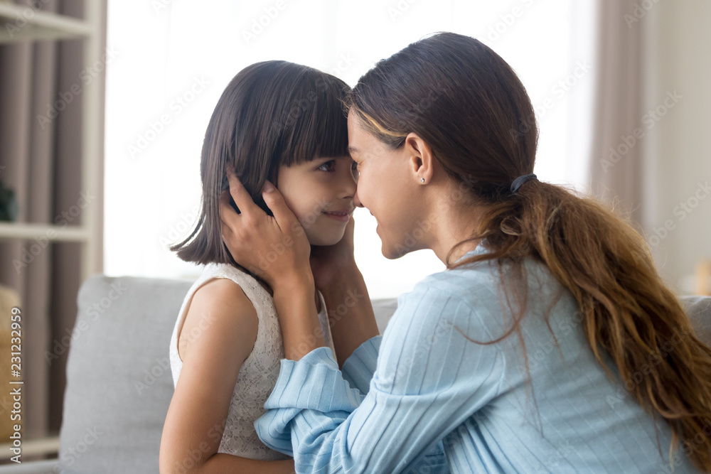 Side view head shot of loving mother and adorable daughter touches with foreheads looking at each other sitting on couch in living room at home. Happy motherhood, new mommy for adopted child concept Side view head shot of loving mother and adorable daughter touches with foreheads looking at each other sitting on couch in living room at home. Happy motherhood, new mommy for adopted child concept