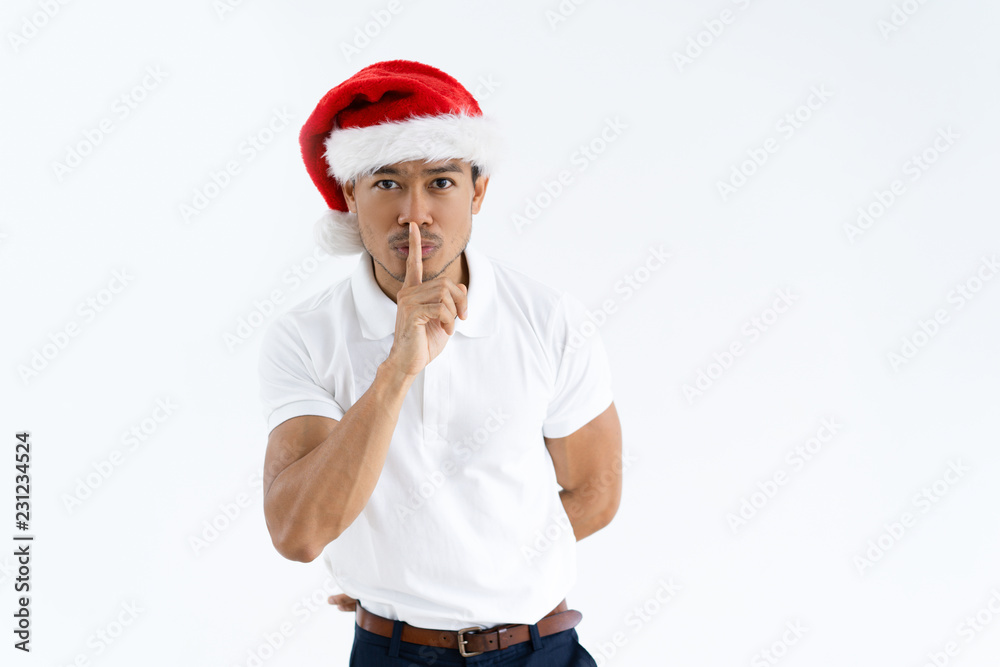 Serious Asian man wearing Santa hat and making silence gesture. Handsome guy touching mouth with forefinger and looking at camera. Christmas surprise concept. Isolated front view on white background. Serious Asian man wearing Santa hat and making silence gesture. Handsome guy touching mouth with forefinger and looking at camera. Christmas surprise concept. Isolated front view on white background.