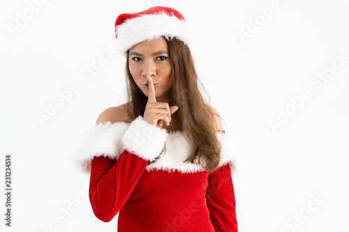 Positive woman wearing Santa hat and making silence gesture. Beautiful Asian lady touching mouth with forefinger and looking at camera. Christmas surprise concept. Isolated view on white background.