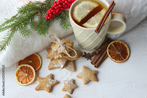 A Cup of tea with lemon on the table close-up surrounded by Christmas decorations and homemade cakes. Star shaped gingerbread, cinnamon sticks and dried oranges on white background.