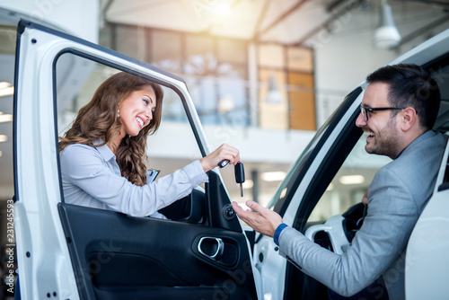 Professional car salesperson handing over keys to the customer of brand new vehicle at local car dealership.