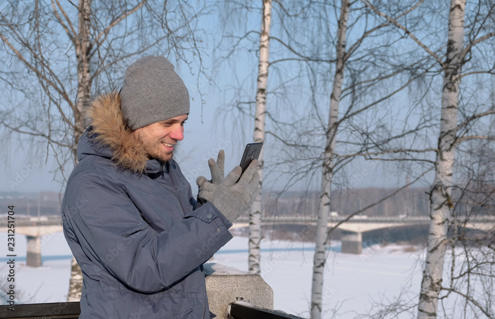 Man in blue down jacket with fur hood using cellphone for video chatting and smiling in winter Park. Man in blue down jacket with fur hood using cellphone for video chatting and smiling in winter Park.