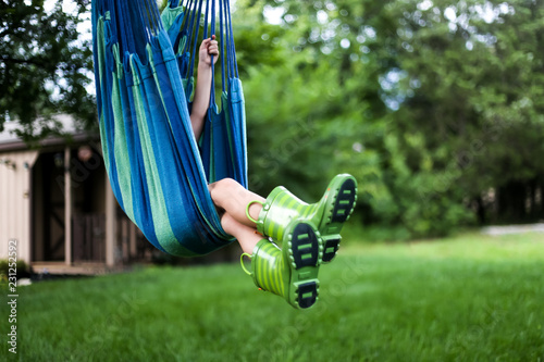 Little Boy in Green Rainboots Swinging Playfully on Tree Hammock Swing with Legs Crossed, Hidden Face, Outdoor Childhood Fun in Nature