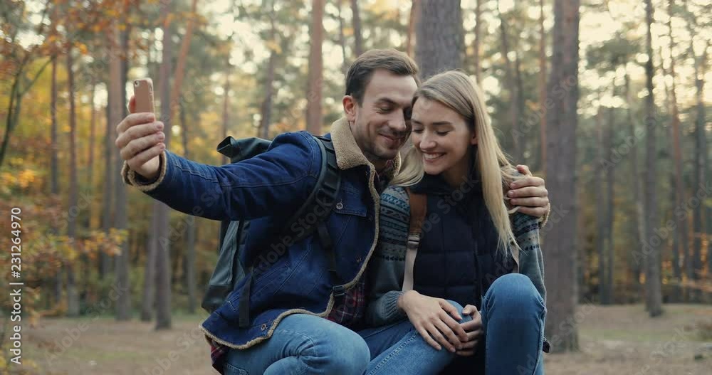 Cheerful young Caucasian couple of tourists sitting in the middle of the forest and taking selfie photo on the smartphone camera. Portrait shot. Outdoors. Cheerful young Caucasian couple of tourists sitting in the middle of the forest and taking selfie photo on the smartphone camera. Portrait shot. Outdoors.