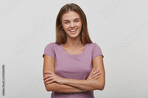 Cheerful young female with pleasant smile, has long brown hair, healthy skin, wears casual t-shirt, stands with hands crossed against white background, being in good mood, stays in a good company