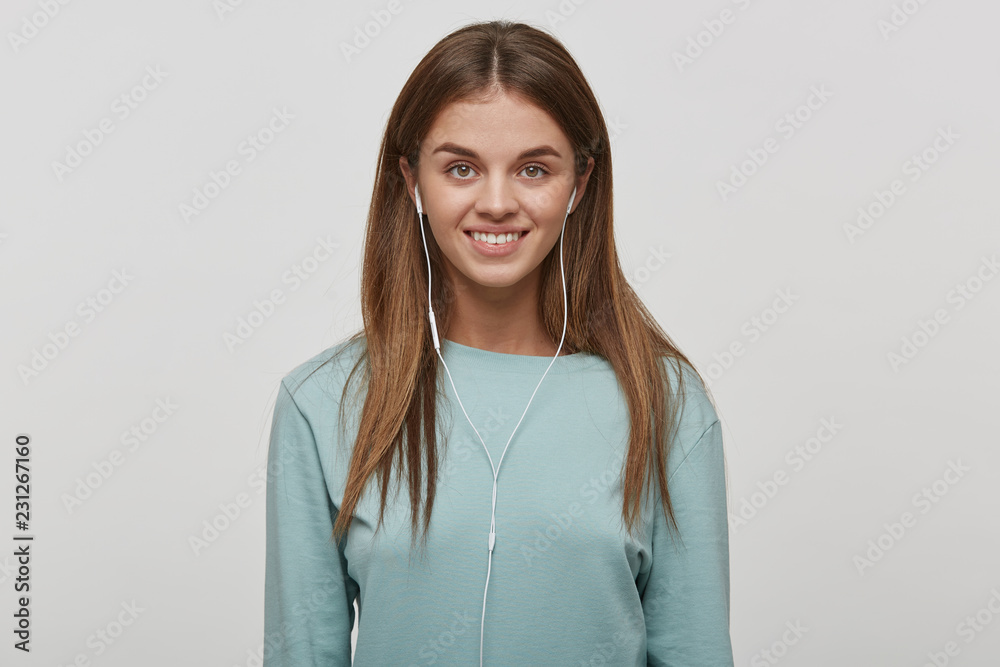 Obraz premium Shoot of a beautiful happy young girl with natural make-up and well-groomed hair, smiles, listens favorite music with headphones, dressed casual, on a white background in the studio.
