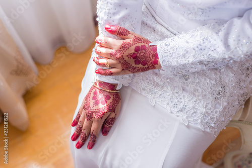 Woman wearing white dress sitting and showing both hands on her lap with henna decoration for wedding. Natural light from window. Blur background