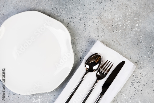 Empty wgite plate, fork, knife, spoon on concrete gray background, top view