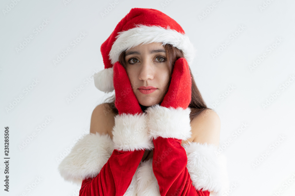 Pensive girl in Santa hat concerned about her face skin. Pretty young woman in Xmas costume touching cheeks with hands in mittens. Celebration concept Pensive girl in Santa hat concerned about her face skin. Pretty young woman in Xmas costume touching cheeks with hands in mittens. Celebration concept