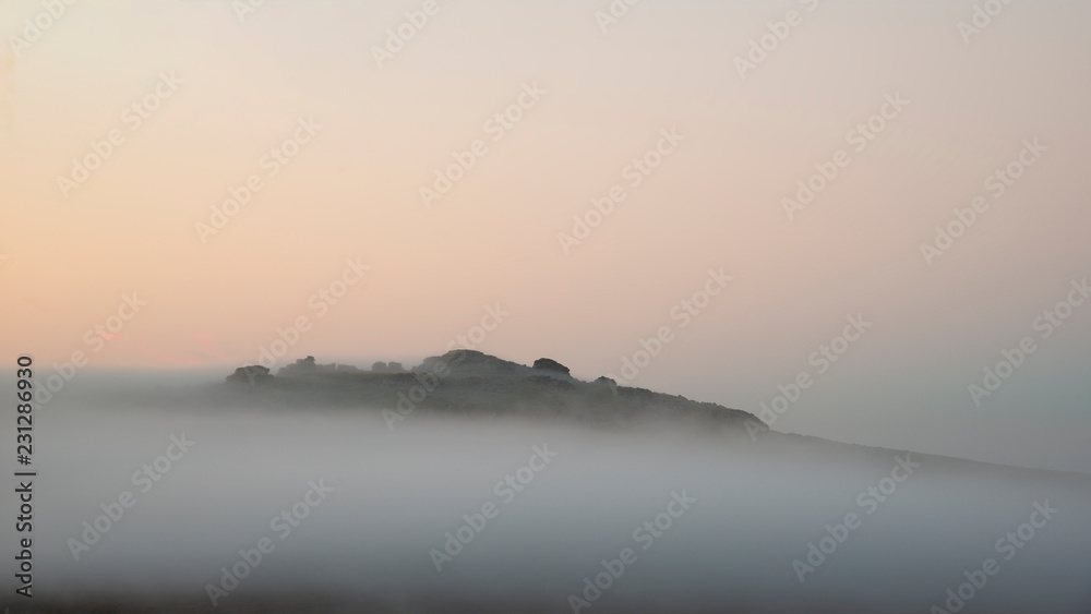 Fototapeta premium Beautiful foggy sunrise landscape over the tors in Dartmoor revealing peaks through the mist