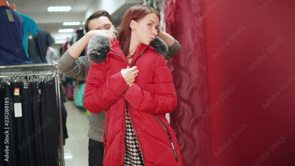 Young guy and his girlfriend in a mall trying out the outerwear in fitting room. Consumerism shopaholism concept. Young guy and his girlfriend in a mall trying out the outerwear in fitting room. Consumerism shopaholism concept.