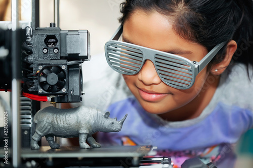Cute girl with cool 3d printed shutter shades is watching her 3d printer as it prints her 3d model of a rhinoceros.