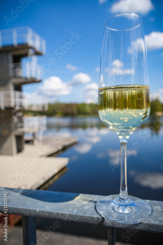 Close-up on a glass of sparkling champagne mirroring the diving platform, blue sky and clouds and the lake in the background
