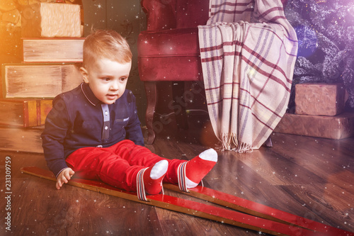 cute interested infant boy wearing ski sitting on the wooden floor near Christmas decorations and big old books