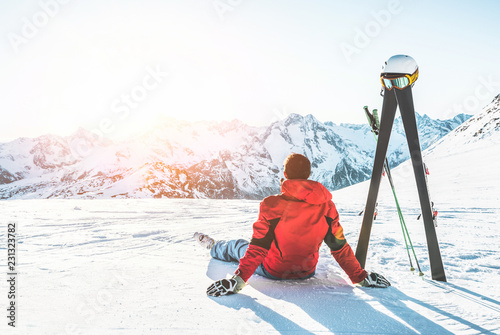 Skier athlete sitting in alpes mountains on sunny day - Adult man enjoying the sunset with skies gear next to him - Winter sport and vacation concept